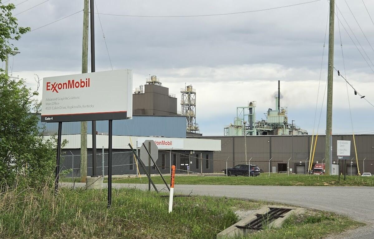 ExxonMobil industrial facility with a large roadside sign in the foreground, gate area, and factory buildings in the background.