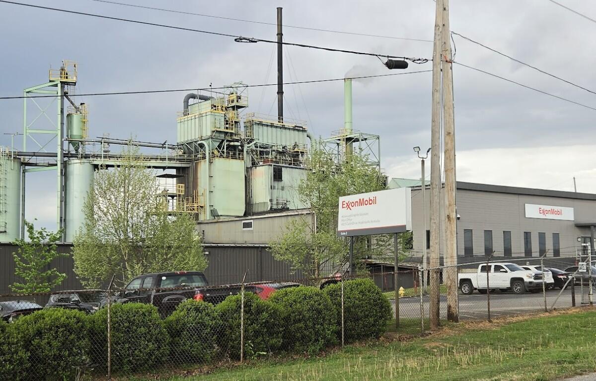 Industrial facility with ExxonMobil sign, large green processing buildings, and a chain-link fence in foreground under cloudy sky