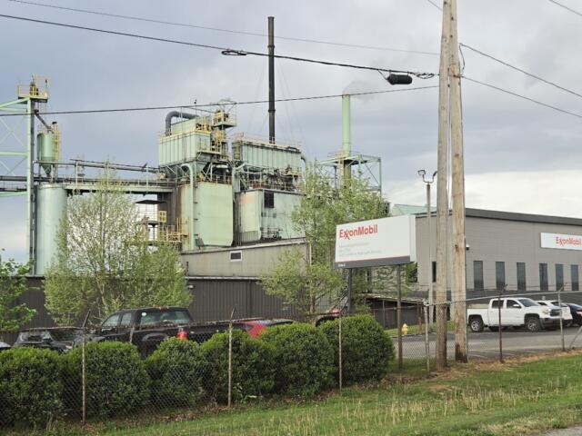 Industrial facility with ExxonMobil sign, large green processing buildings, and a chain-link fence in foreground under cloudy sky