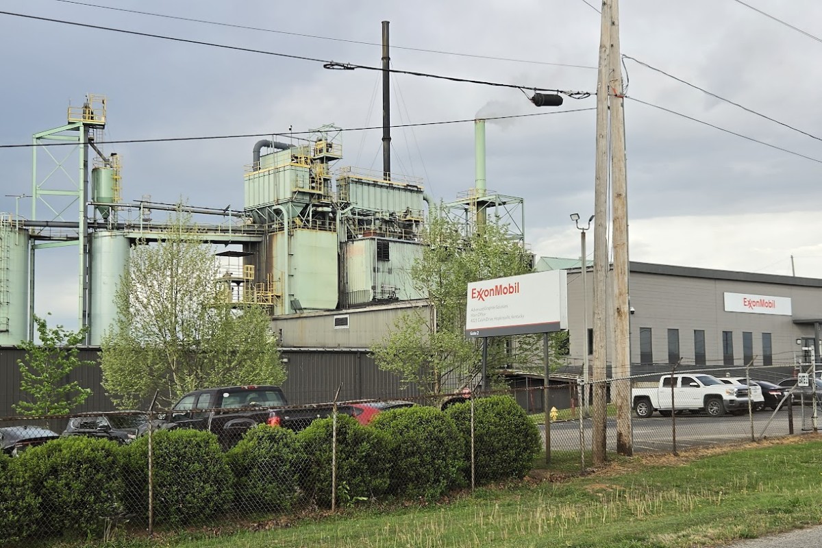Industrial facility with ExxonMobil sign, large green processing buildings, and a chain-link fence in foreground under cloudy sky