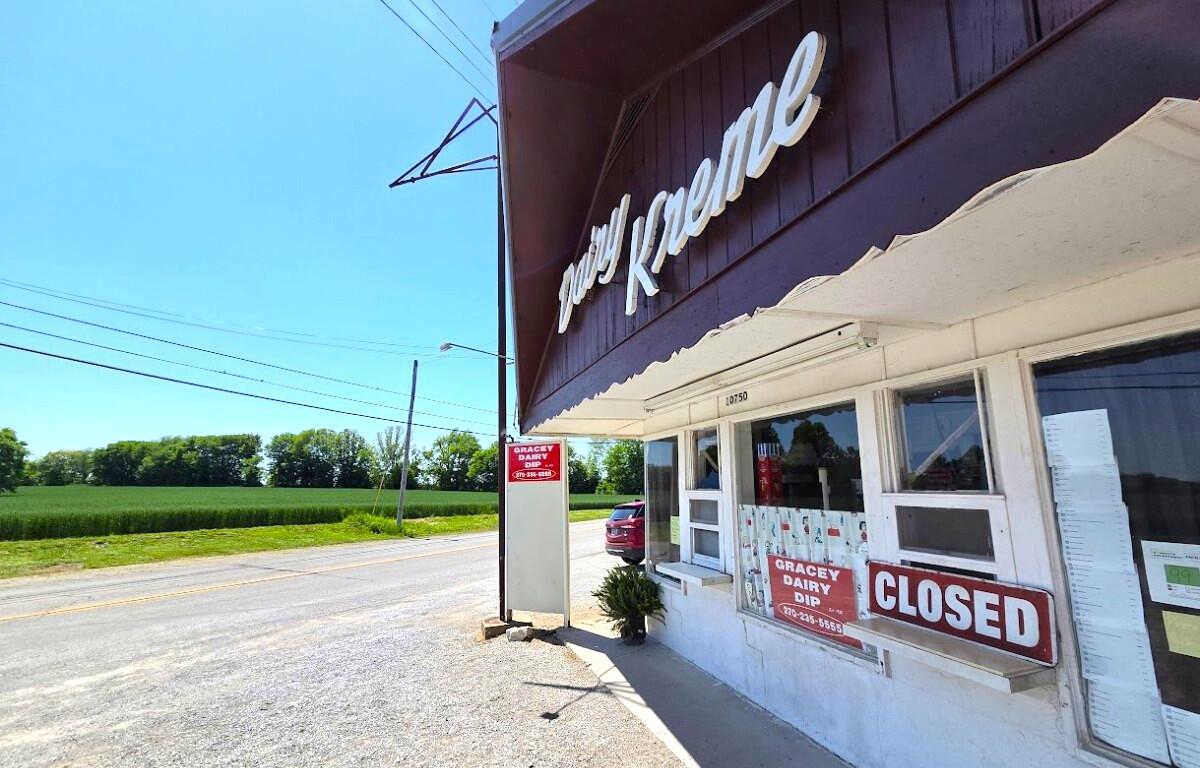 Small rural storefront with a large 'CLOSED' sign in the window, beside a road under a bright blue sky.