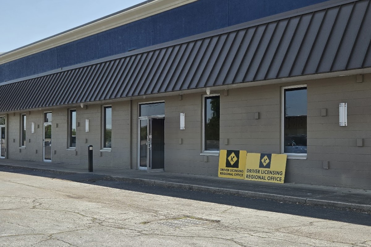 Exterior view of a beige office building with a blue upper trim and diagonal metal awning, near two Driver Licensing Regional Office signs outside the entrance.
