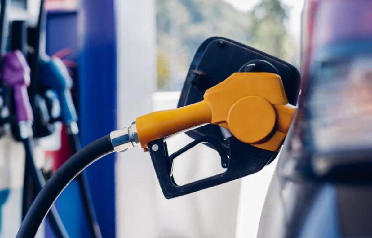 Orange fuel nozzle inserted into a car's fuel port at a gas station, hoses connected to the tank in the foreground.
