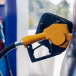 Orange fuel nozzle inserted into a car's fuel port at a gas station, hoses connected to the tank in the foreground.