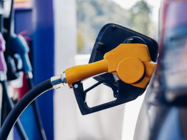Orange fuel nozzle inserted into a car's fuel port at a gas station, hoses connected to the tank in the foreground.