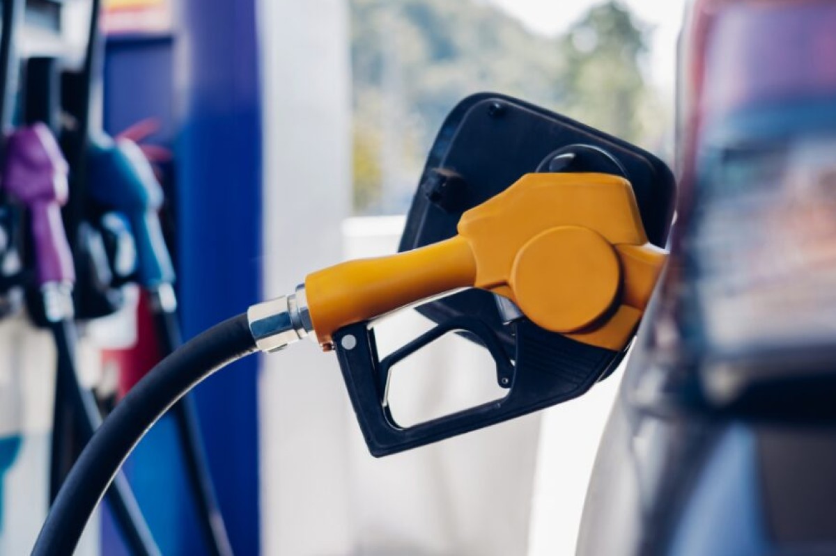 Orange fuel nozzle inserted into a car's fuel port at a gas station, hoses connected to the tank in the foreground.