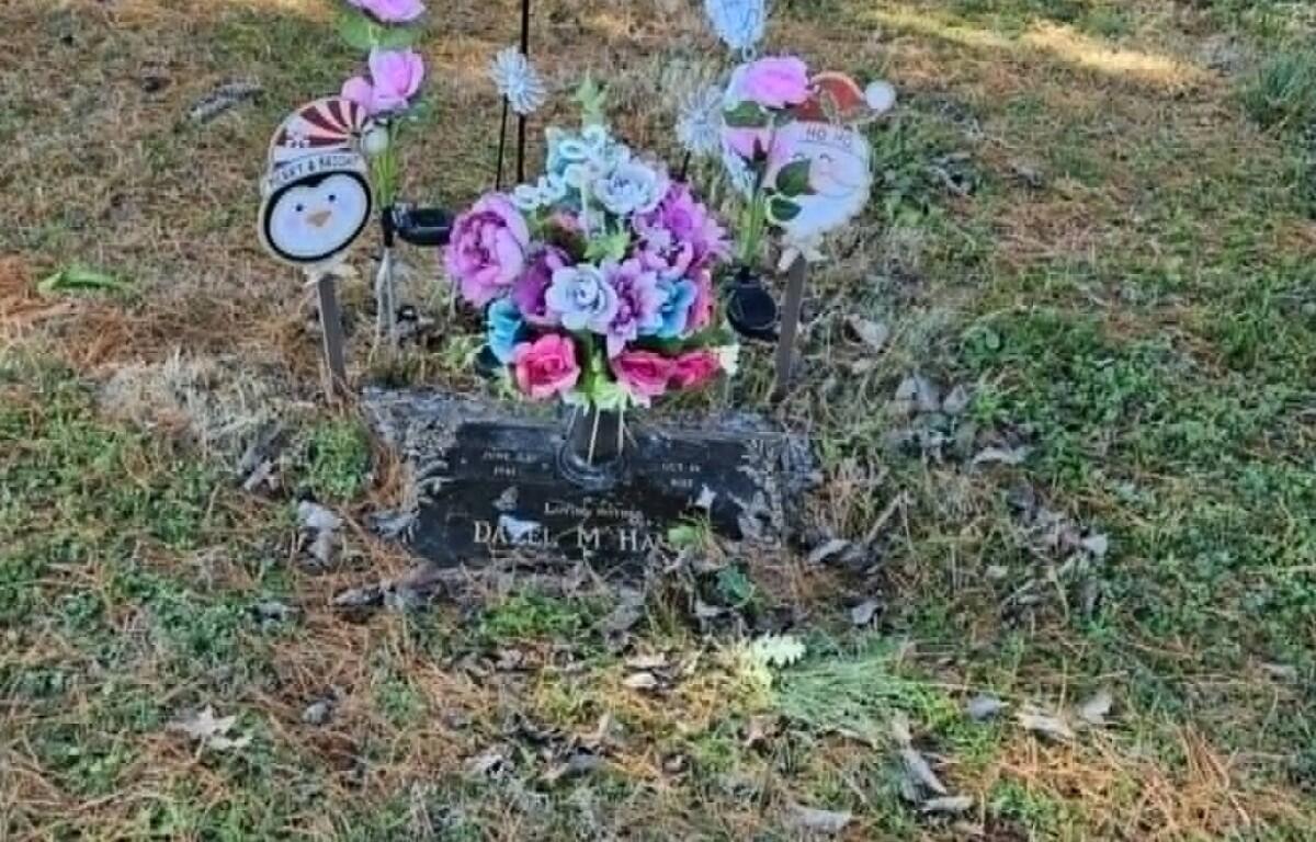 Grave marker with a colorful bouquet of artificial flowers in a vase, surrounded by small decorations in a grassy cemetery.
