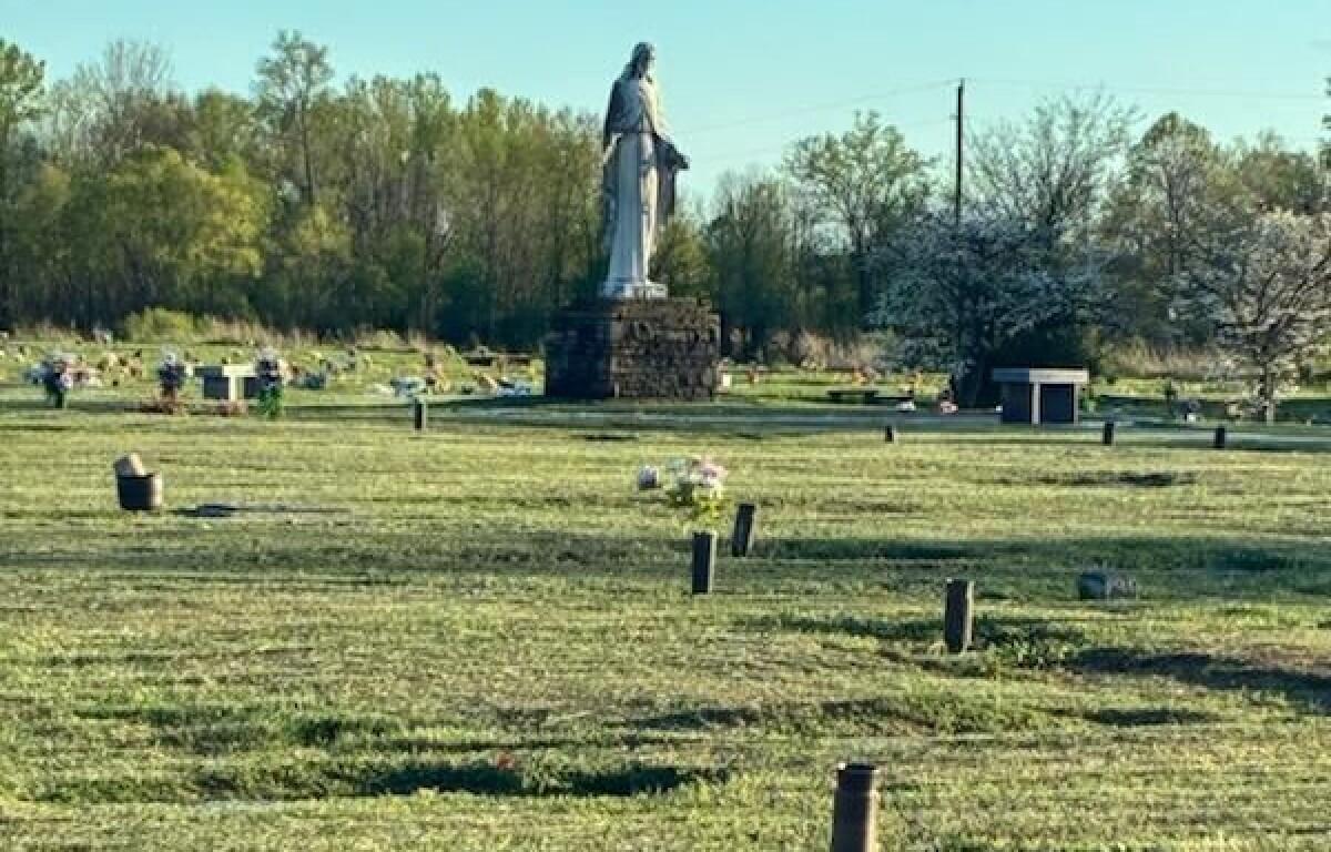 A cemetery scene with a tall statue on a stone pedestal in the center and rows of headstones across a grassy field, trees in the background.