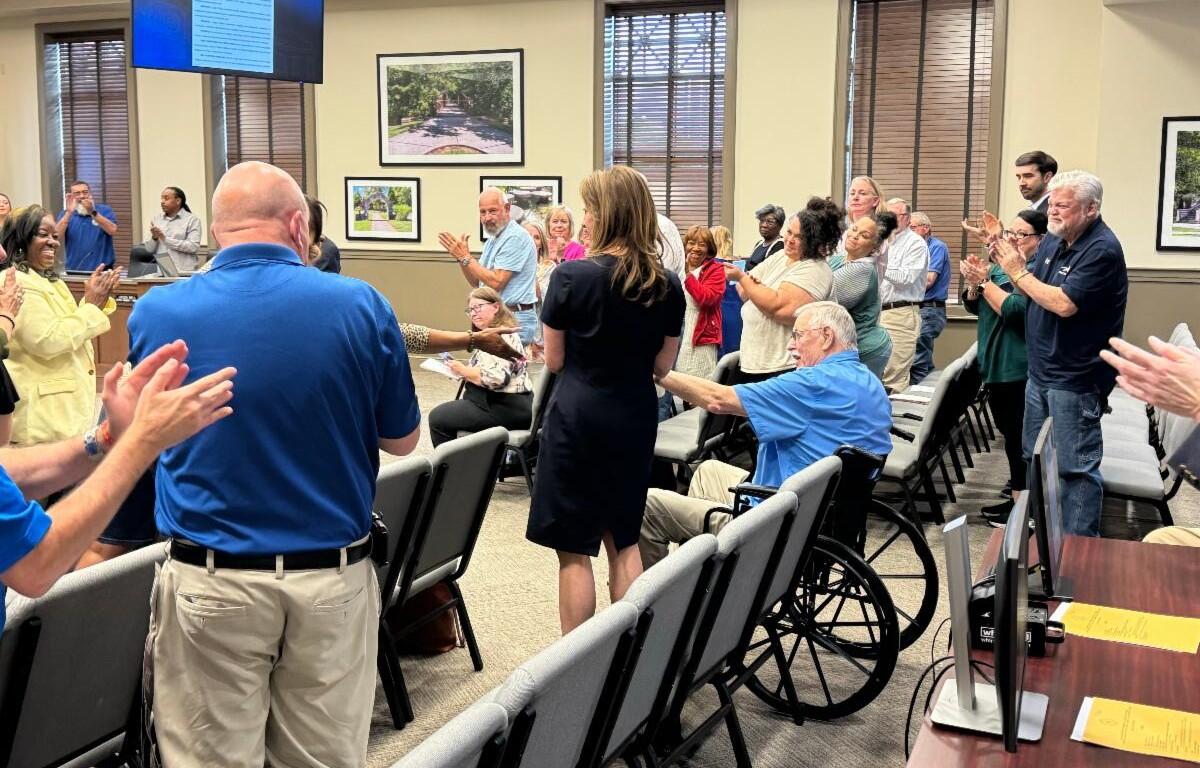 Audience in a meeting room applaud around a central woman in a black dress; a man in a wheelchair and others seated or standing present.
