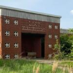 Abandoned red brick building with square decorative blocks and a dark open entrance, surrounded by tall grasses and shrubs in front.