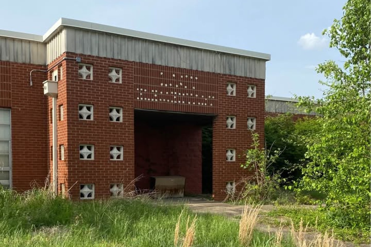 Abandoned red brick building with square decorative blocks and a dark open entrance, surrounded by tall grasses and shrubs in front.