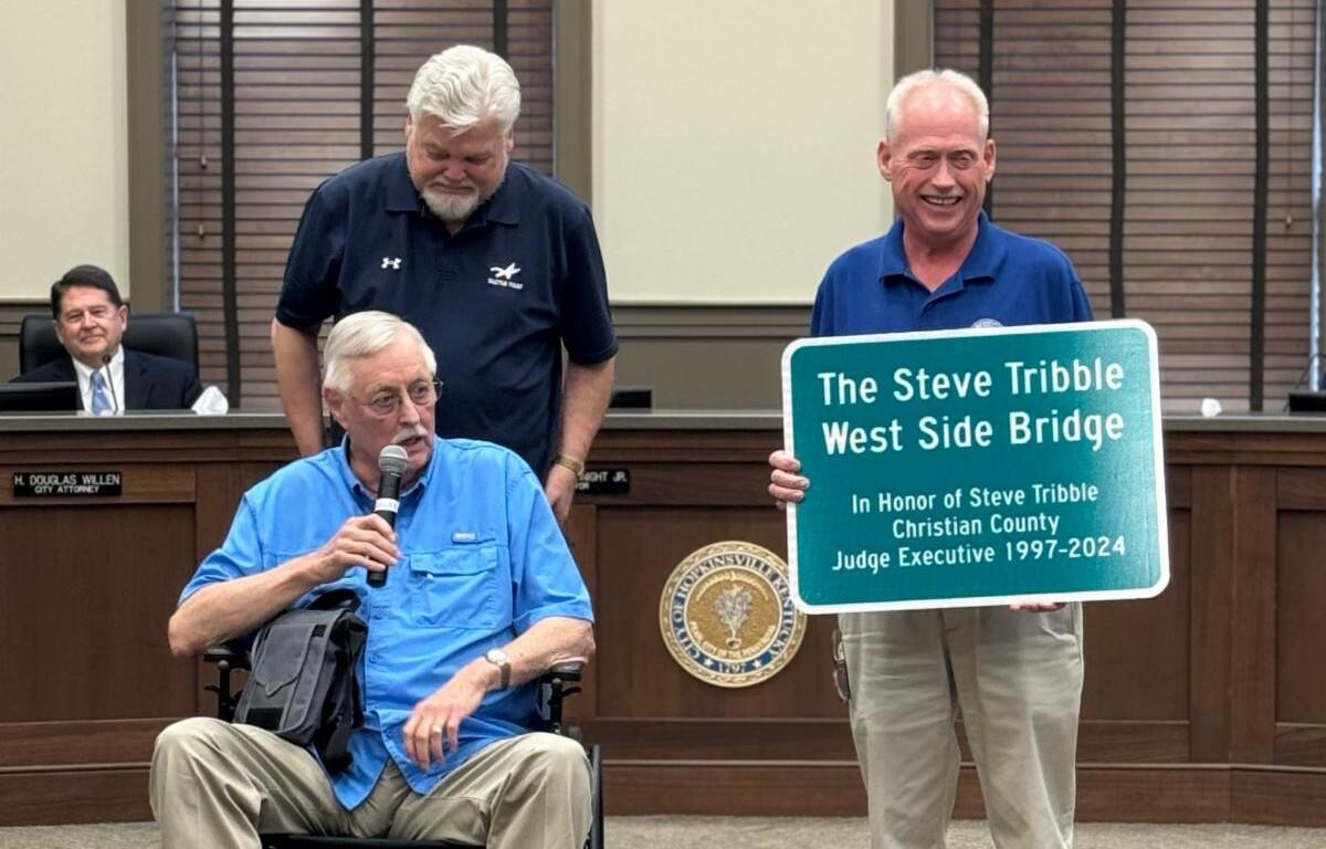 Man in a blue shirt in a wheelchair speaks into a microphone while two men stand nearby, one holding a sign that reads 'The Steve Tribble West Side Bridge' in a government meeting room.
