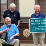 Man in a blue shirt in a wheelchair speaks into a microphone while two men stand nearby, one holding a sign that reads 'The Steve Tribble West Side Bridge' in a government meeting room.