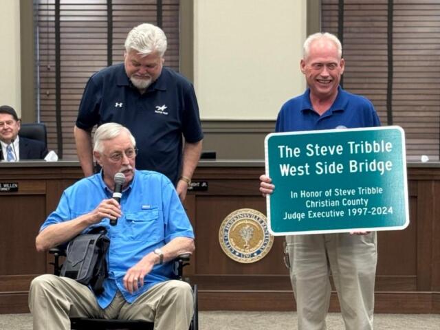 Man in a blue shirt in a wheelchair speaks into a microphone while two men stand nearby, one holding a sign that reads 'The Steve Tribble West Side Bridge' in a government meeting room.