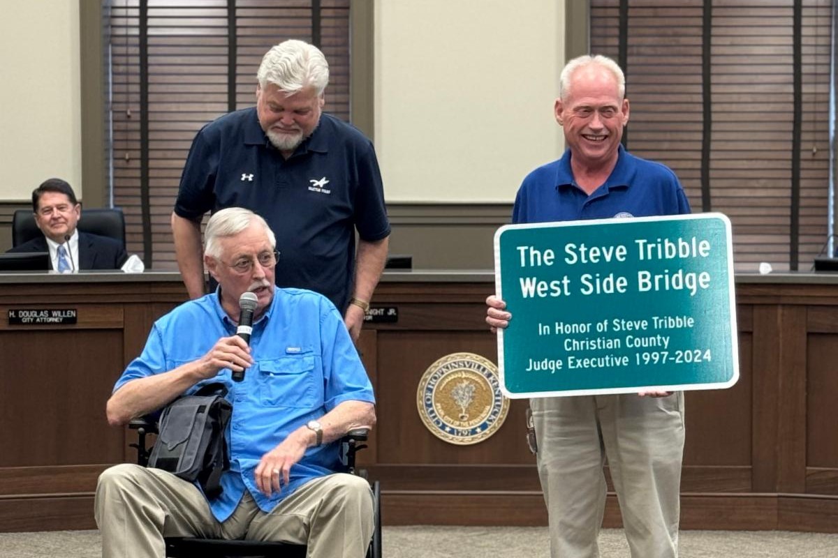 Man in a blue shirt in a wheelchair speaks into a microphone while two men stand nearby, one holding a sign that reads 'The Steve Tribble West Side Bridge' in a government meeting room.