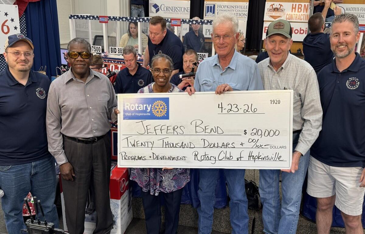 Eight adults stand on a stage, holding a large ceremonial check made out to Jeffers Bend for $20,000 dated 4-23-26, from the Rotary Club of Hopkinsville, at a fundraiser event, with banners and volunteers in the background.