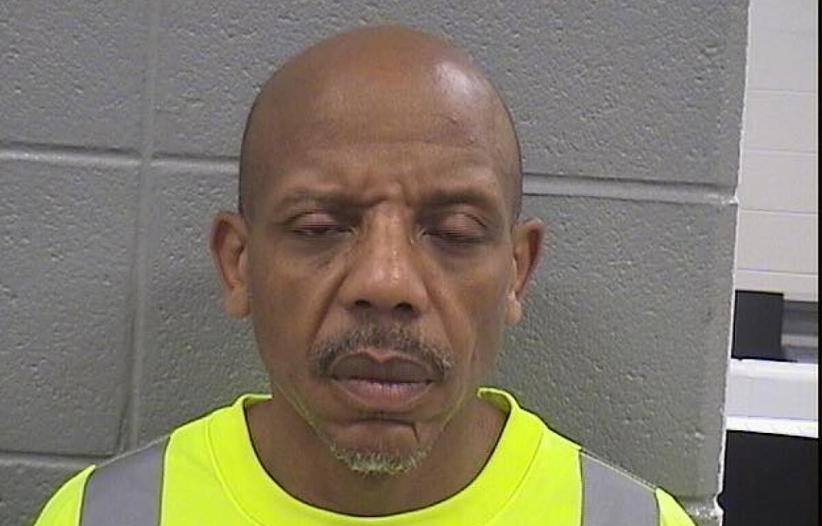 Man with a shaved head and goatee wearing a bright neon safety vest, standing against a gray cinder block wall, eyes closed and mouth slightly open.
