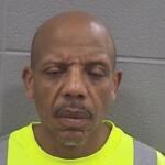 Man with a shaved head and goatee wearing a bright neon safety vest, standing against a gray cinder block wall, eyes closed and mouth slightly open.