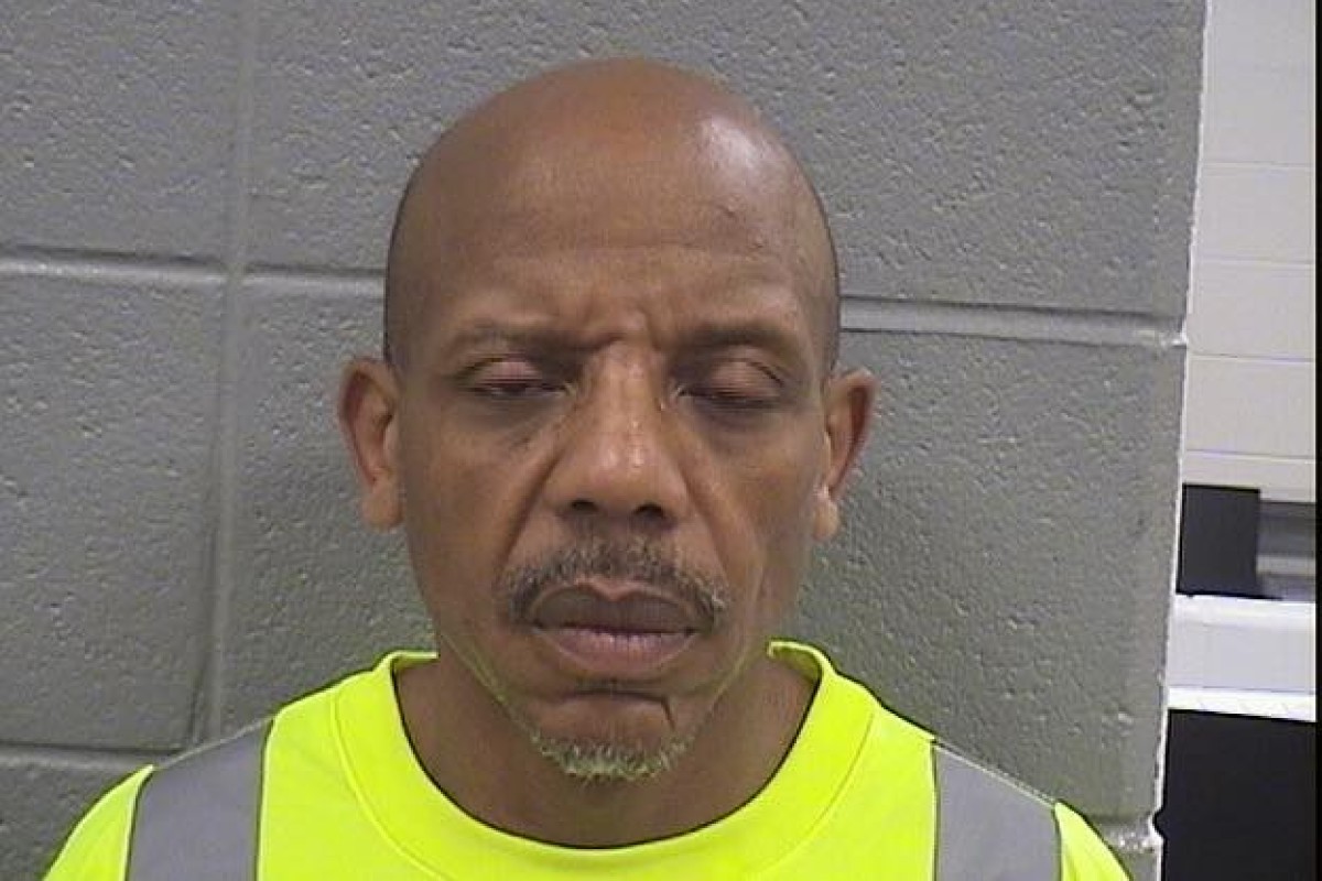 Man with a shaved head and goatee wearing a bright neon safety vest, standing against a gray cinder block wall, eyes closed and mouth slightly open.