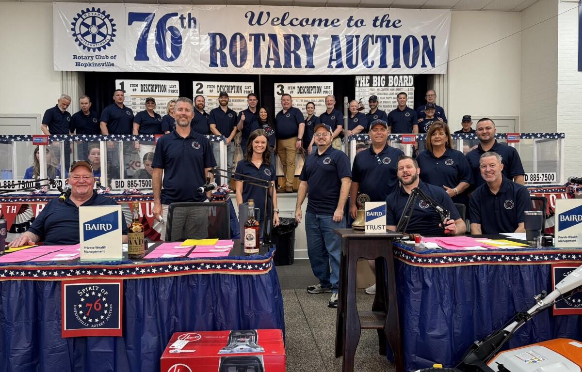 Group of Rotary Club members posing on stage at a fundraising auction, with a banner reading 'Welcome to the 76th Rotary Auction' in the background