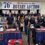 Group of Rotary Club members posing on stage at a fundraising auction, with a banner reading 'Welcome to the 76th Rotary Auction' in the background