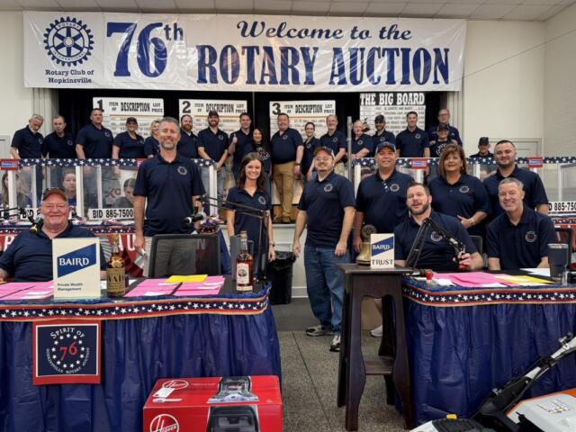 Group of Rotary Club members posing on stage at a fundraising auction, with a banner reading 'Welcome to the 76th Rotary Auction' in the background