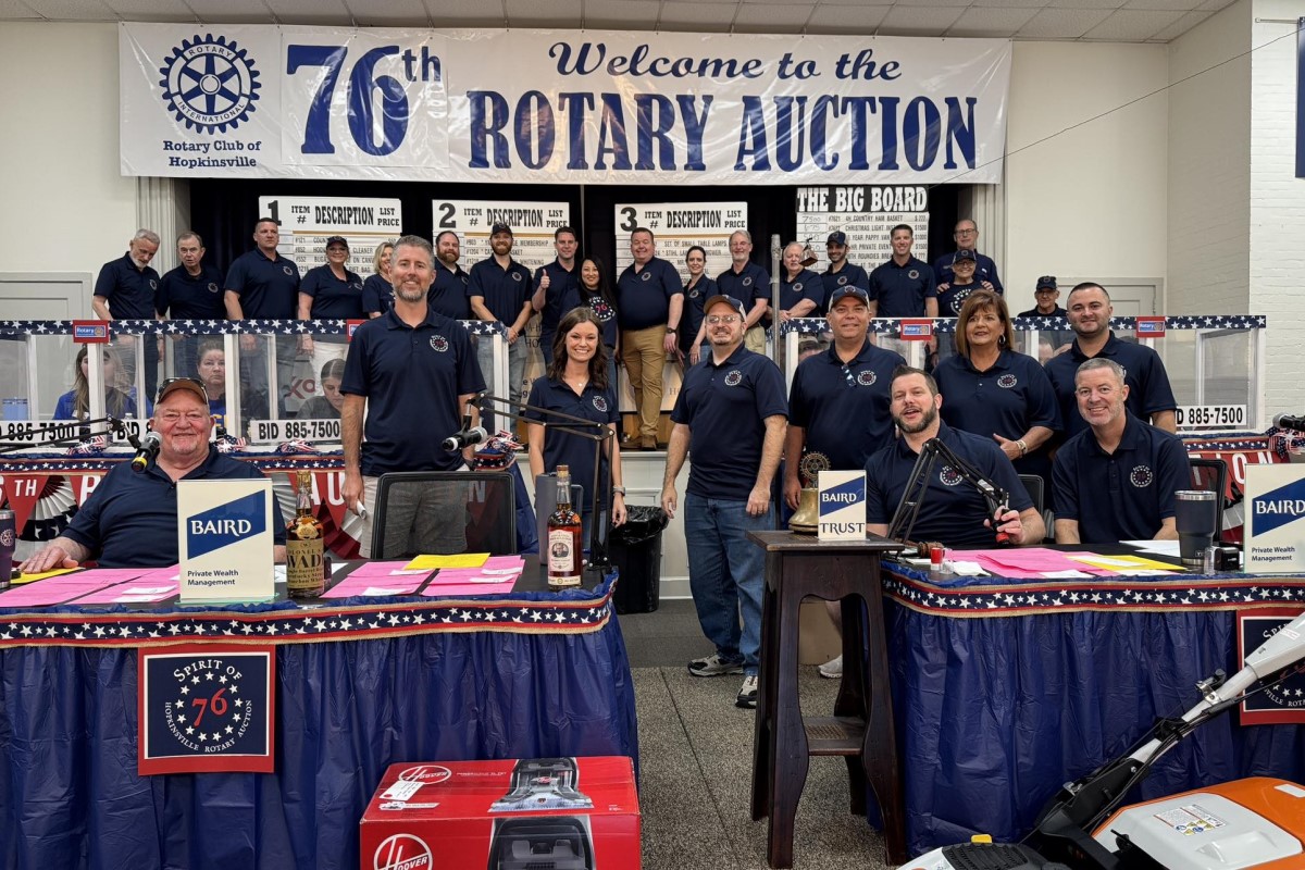Group of Rotary Club members posing on stage at a fundraising auction, with a banner reading 'Welcome to the 76th Rotary Auction' in the background