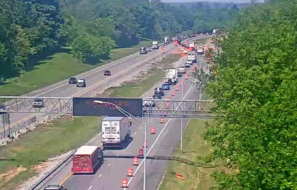 Highway construction zone with multiple lanes closed, orange traffic cones, and trucks; green trees on both sides.