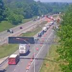 Highway construction zone with multiple lanes closed, orange traffic cones, and trucks; green trees on both sides.