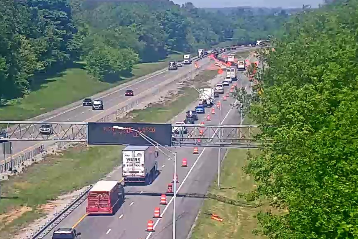 Highway construction zone with multiple lanes closed, orange traffic cones, and trucks; green trees on both sides.