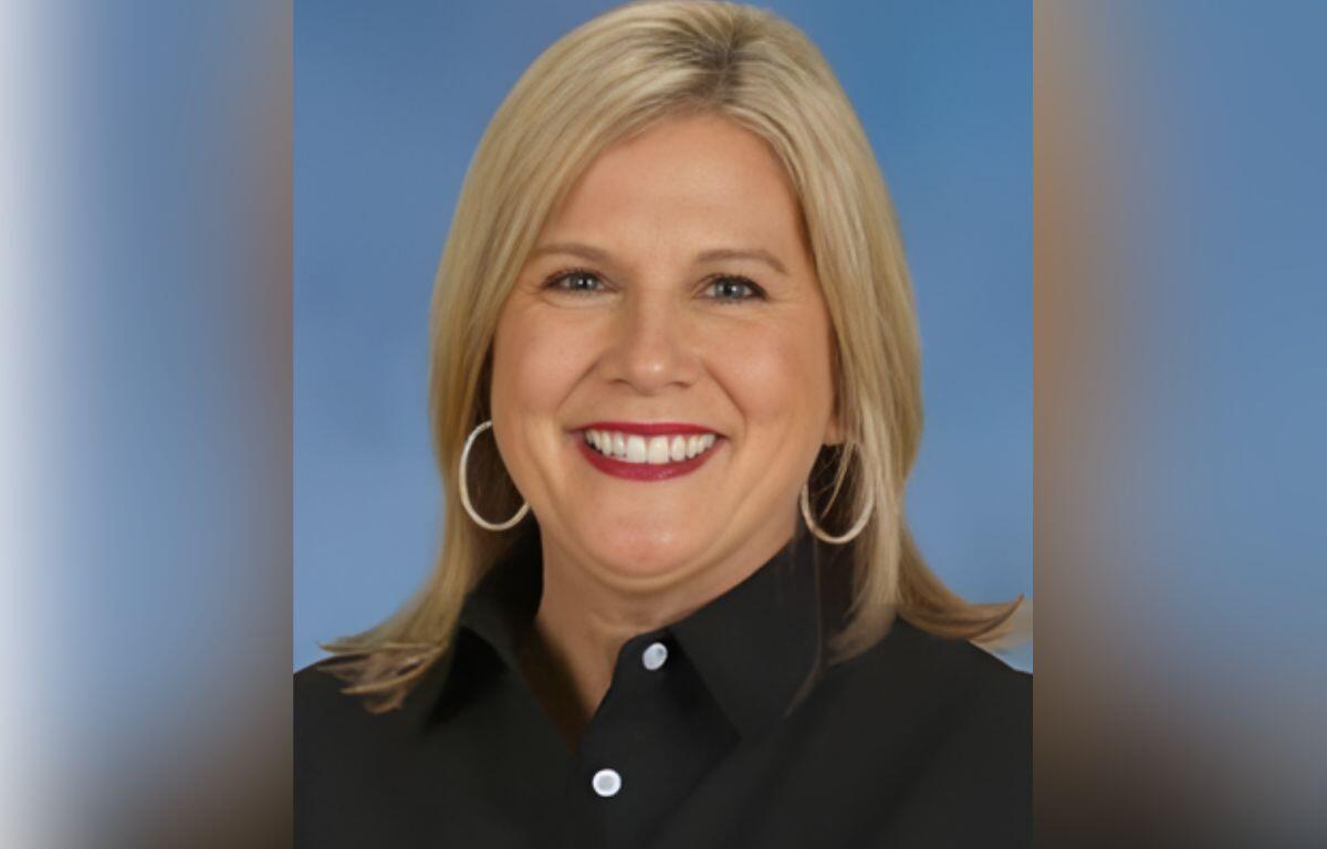 Portrait of a smiling blonde woman wearing a black button-down shirt and hoop earrings against a blue background.