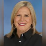 Portrait of a smiling blonde woman wearing a black button-down shirt and hoop earrings against a blue background.