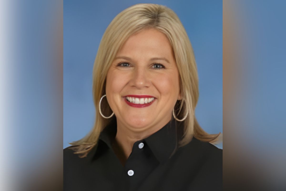 Portrait of a smiling blonde woman wearing a black button-down shirt and hoop earrings against a blue background.