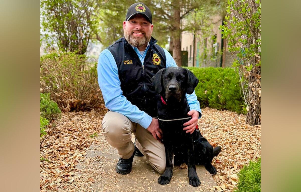 Kneeling man in uniform beside a black Labrador on a leaf-strewn sidewalk outdoors, posing for a photo.