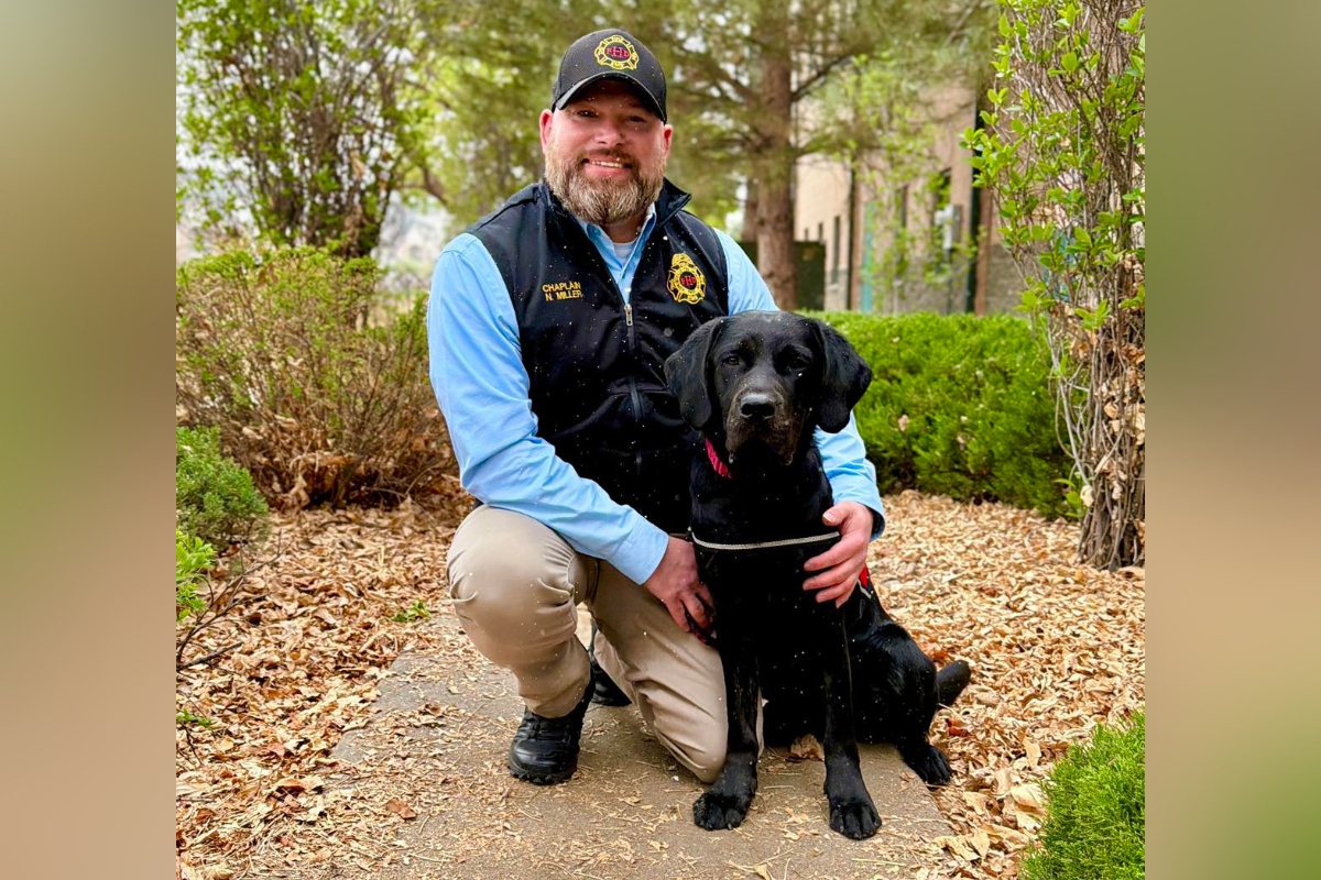 Kneeling man in uniform beside a black Labrador on a leaf-strewn sidewalk outdoors, posing for a photo.