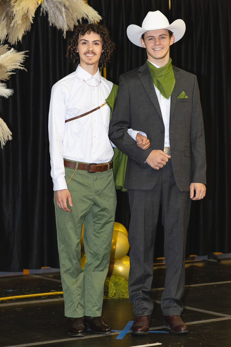 Two young men in western costumes pose arm-in-arm on a stage, one in a white shirt and green pants, the other in a gray suit with a white cowboy hat and green scarf.