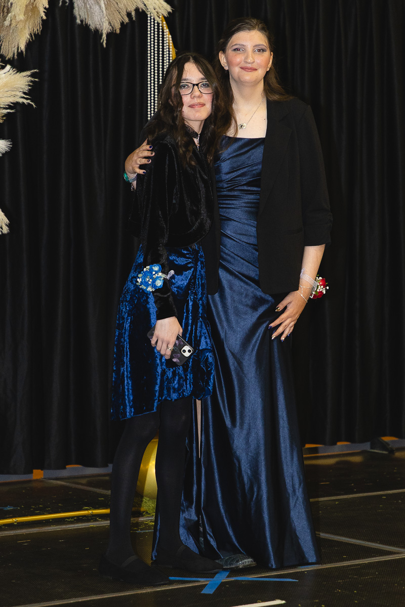 Two young women in formal dresses pose together on a stage, one in blue velvet and the other in a navy satin gown with a black blazer.