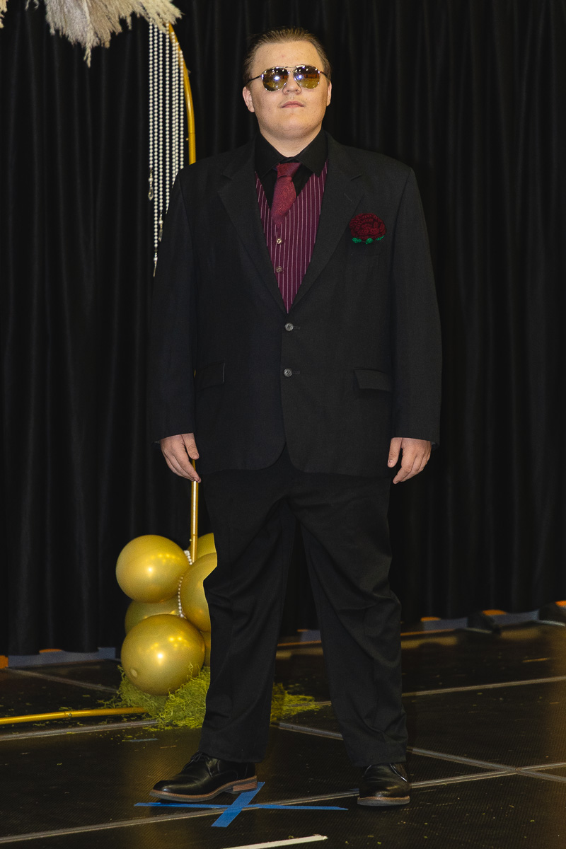 Man in a dark suit with burgundy vest and sunglasses standing on a stage with gold balloons in the background.