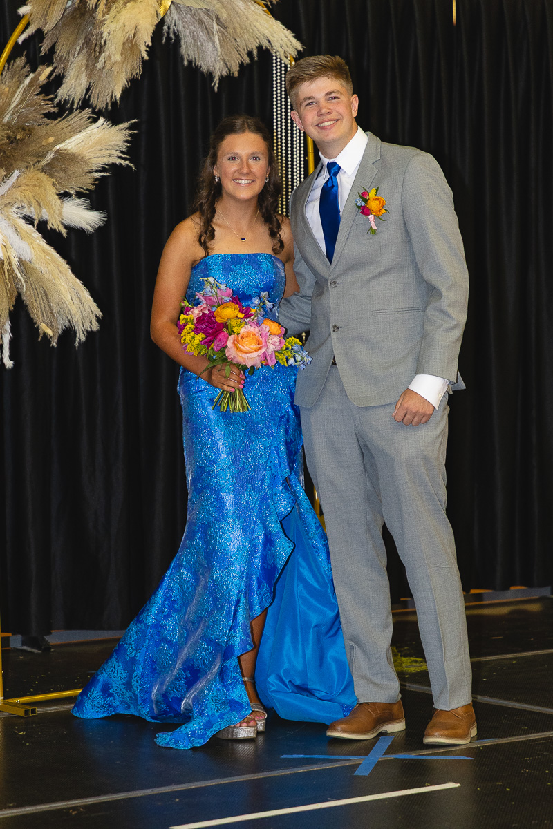 Teen couple posing on a stage in formal attire; girl in a blue sequined gown holding a colorful bouquet, boy in a gray suit with a blue tie and a boutonniere.