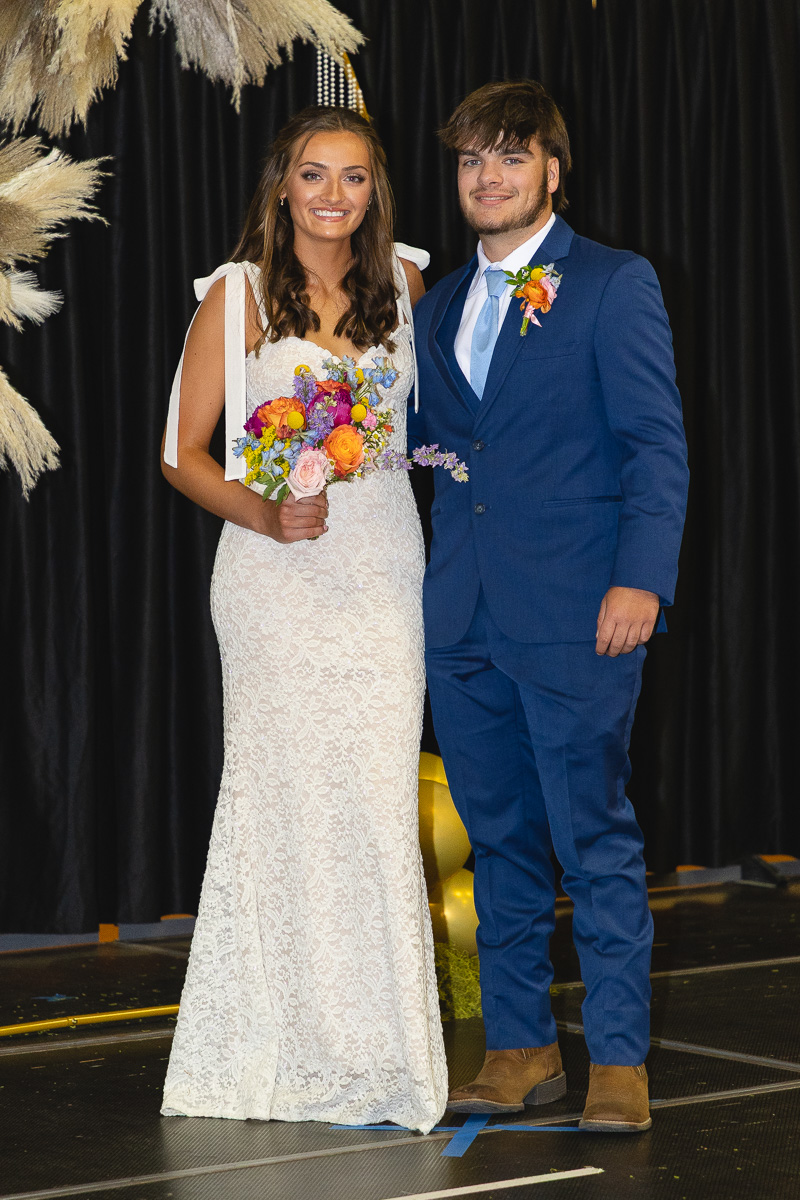 Bride in white lace gown holding a colorful bouquet, standing with a groom in a blue suit on stage with black curtains and pampas decor