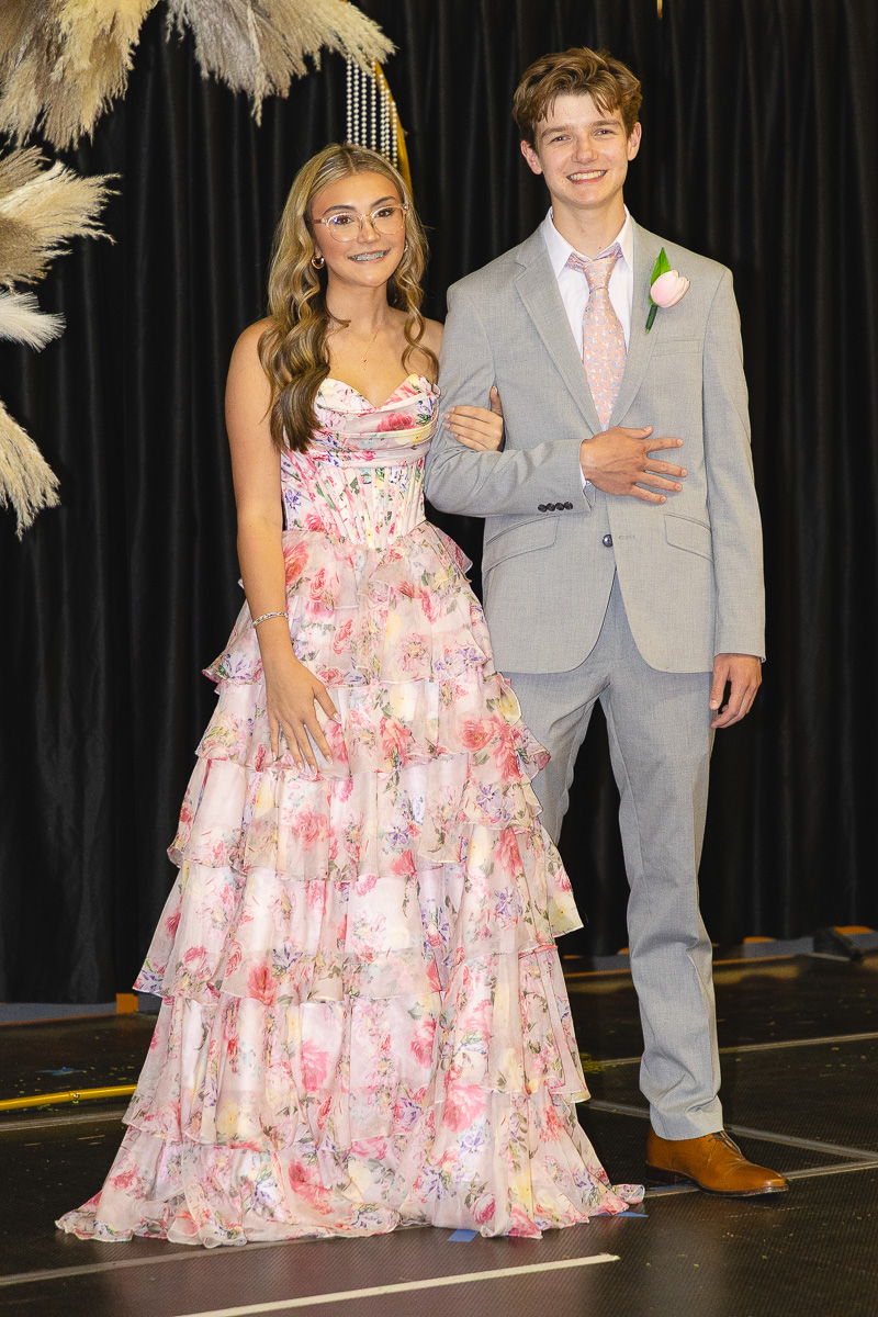 Teen couple posing on stage: girl in a pink floral ball gown with braces and glasses, boy in a light gray suit with a pink tie and boutonniere, arm linked together.