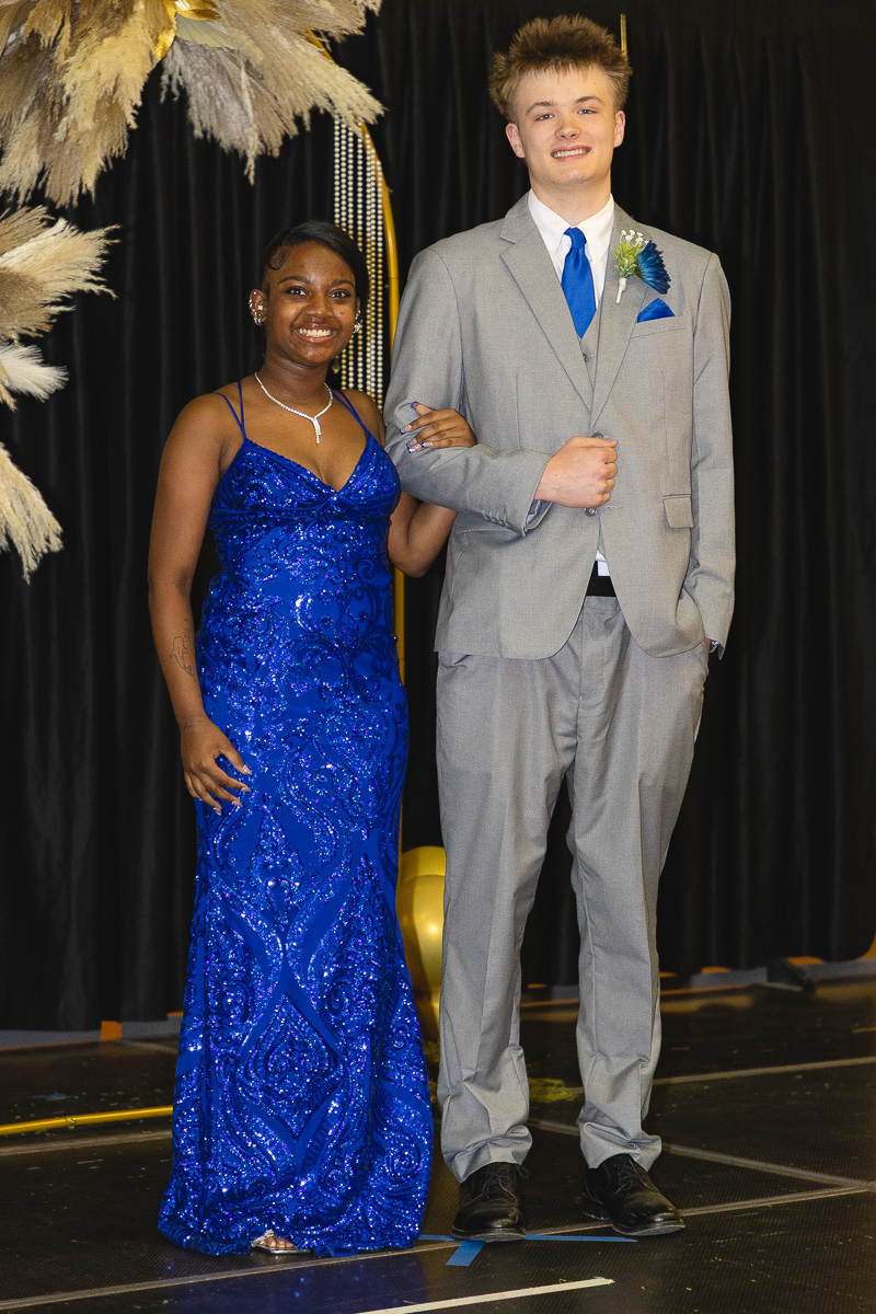 Young woman in a royal blue sequined gown and a young man in a light gray suit with a blue tie pose arm-in-arm on a stage, smiling.