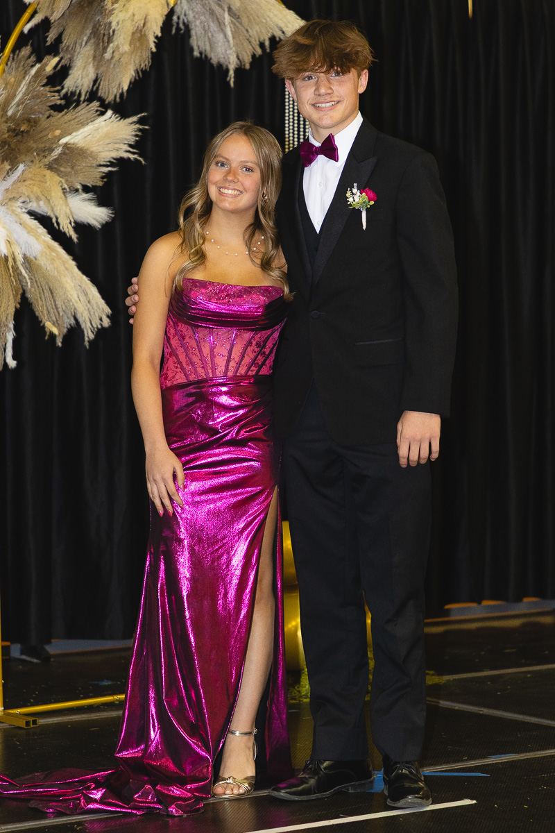 Two teens in formal wear posing together on a stage: girl in a magenta dress with a high slit, boy in a black tuxedo with burgundy bow tie and boutonniere.
