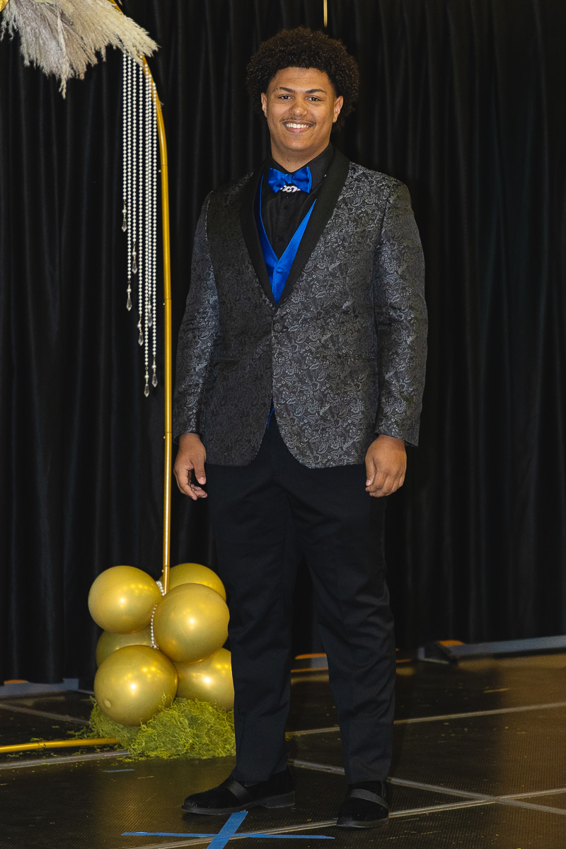Smiling young man in a patterned blazer and blue bow tie posing on a stage with gold balloons in the background at a formal event.