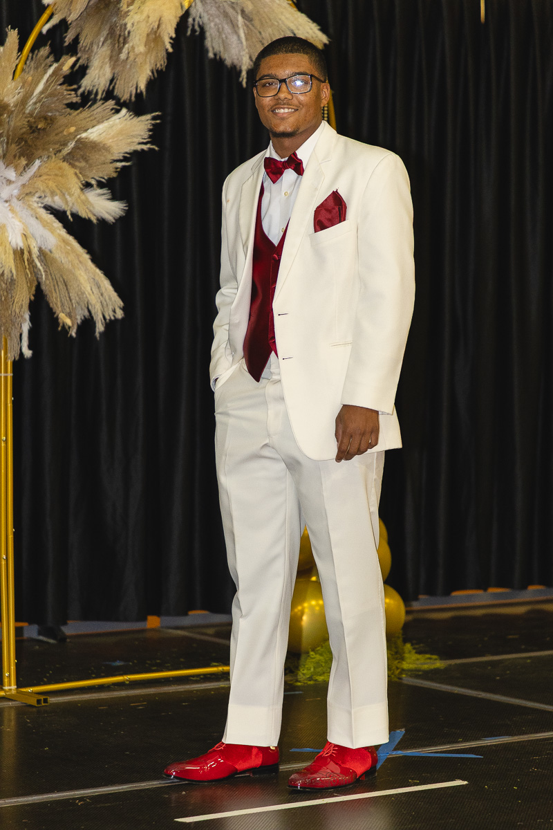 Man in a white suit with red vest, bow tie, and pocket square posing on a stage with black backdrop and gold decorations.
