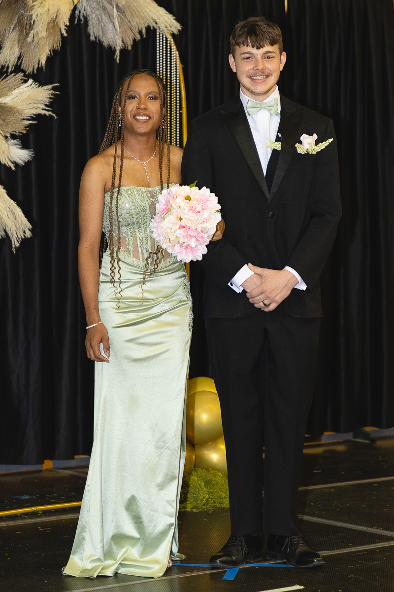 Two teens in formal wear posing on a stage; girl in a mint gown with a pink bouquet, boy in a black suit with a pale green bow tie and boutonniere.”,