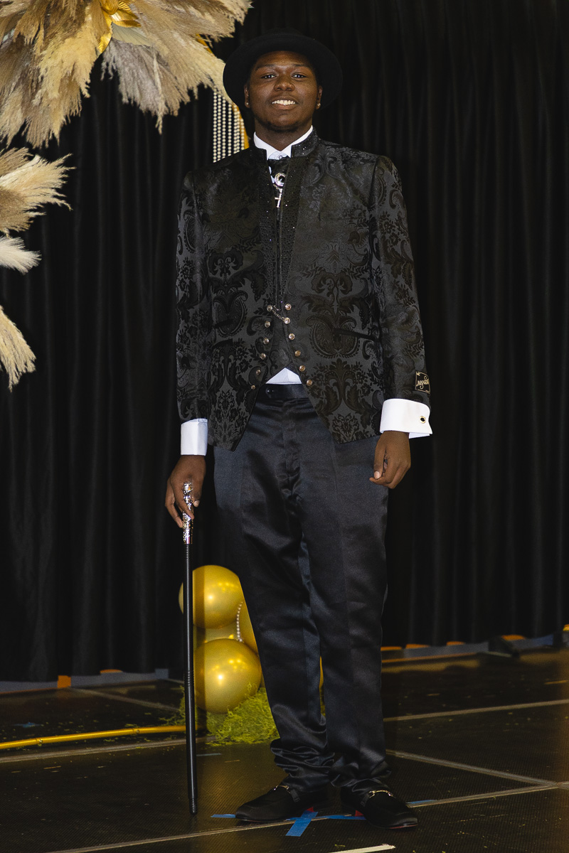 Young man in a black brocade suit with white cuffs and a cane on stage, gold balloons in the background.