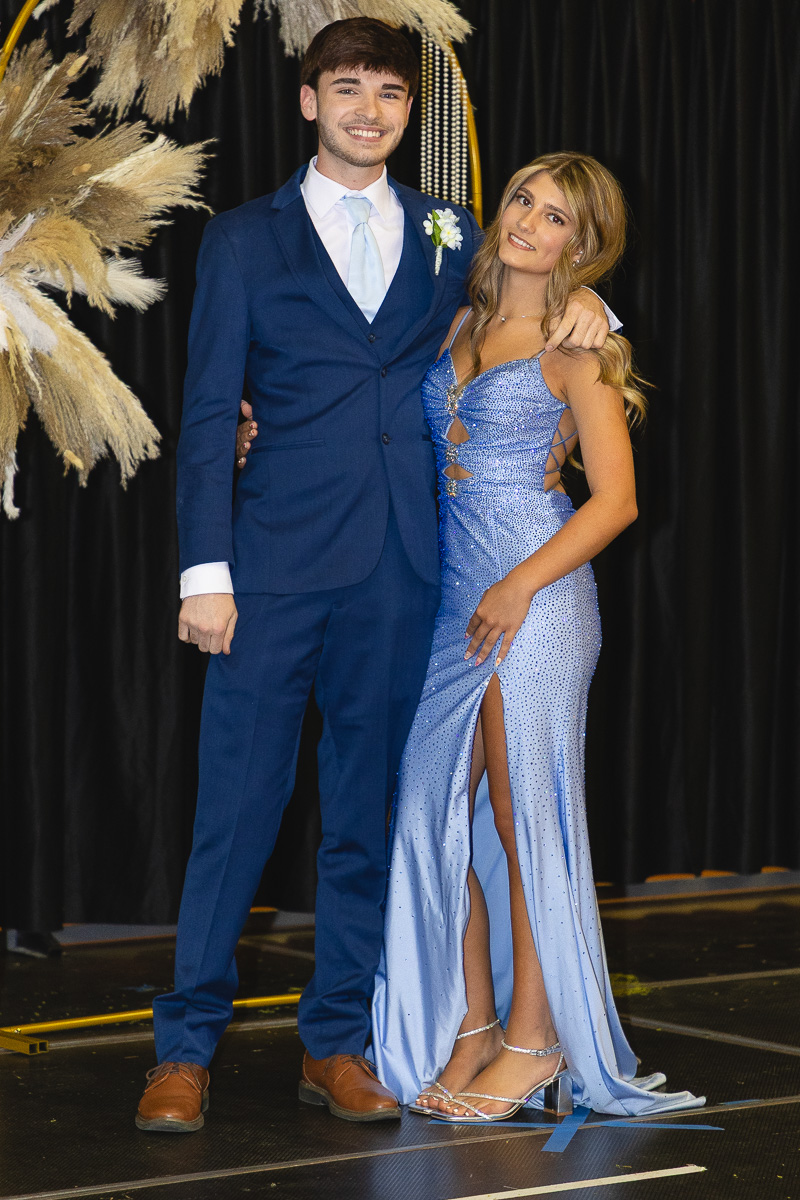 Young couple in formal attire posing arm in arm on a stage with a black curtain backdrop and pampas decorations.