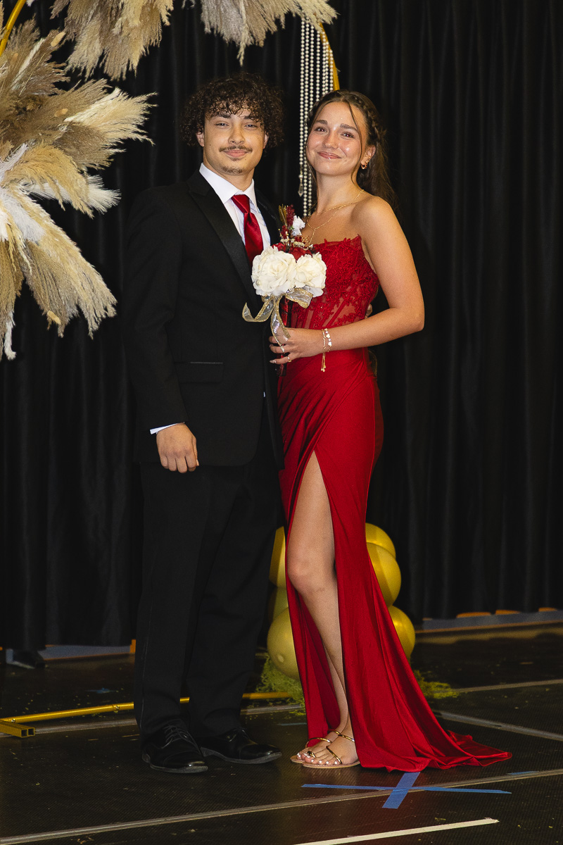 A young man in a black suit and red tie stands beside a woman in a red, high-slit gown holding a bouquet, posing on a stage with decorative pampas grass.