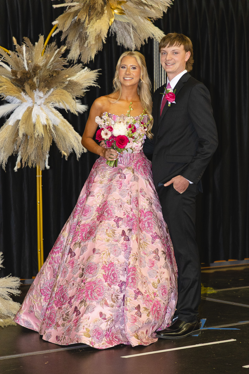 Couple posing in formal attire on a stage; woman in a pink floral ball gown holds a pink bouquet, man in a dark suit with a pink boutonniere stands beside her.